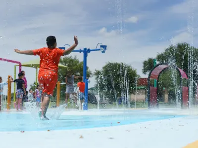 Child in orange shirt and shorts playing at a splash pad while water falls from above.