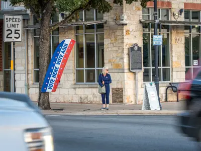 A woman stands on a sidewalk near a polling place sign reading "VOTE HERE - VOTE AQUÍ," with cars passing by on the street and a speed limit 25 sign visible.