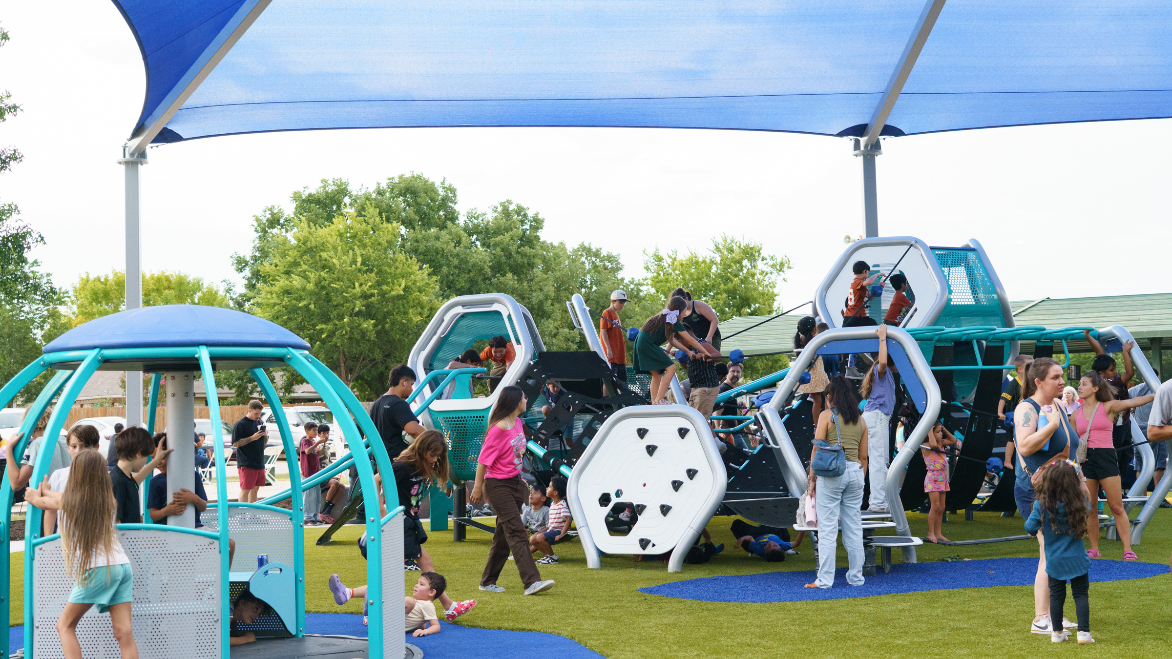 Children play at a new playground at Steeplechase Park in Kyle, TX. The playground has a bright green turf with blue play features. 