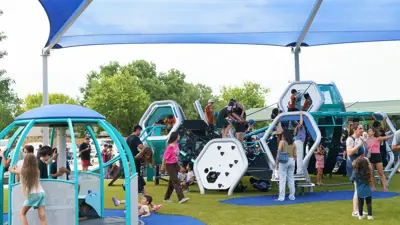 Children play at a new playground at Steeplechase Park in Kyle, TX. The playground has a bright green turf with blue play features.