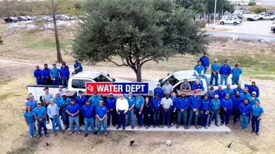 A photograph of the City of Kyle Water Utilities Department all standing together, wearing predominantly blue button up shirts gathering around a light blue truck and a sign that says 'KYLE.'
