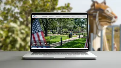 A computer sits on a table with 'City of Kyle Annual Budget' written in the lower left hand corner. In the background is a wooden piecemeal bobcat that is part of a local playground in Kyle, TX.