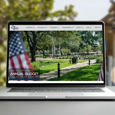 A computer sits on a table with 'City of Kyle Annual Budget' written in the lower left hand corner. In the background is a wooden piecemeal bobcat that is part of a local playground in Kyle, TX.