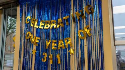 Blue streamers hang against a wall that states 'Celebrating one year of 3-1-1' in gold lettering. 