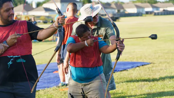 A young man wearing a red jersey holds a bow and arrow while being instructed by a teacher. 