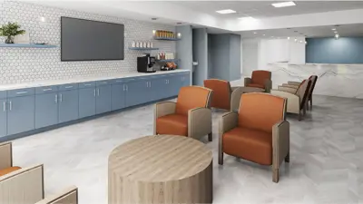 A bright lounge area with blue cabinetry, a coffee station, wall-mounted TV, and cushioned chairs arranged around small tables for casual seating.