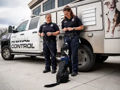 Two animal control officers stand beside a truck labeled "Kyle Animal Control," smiling at a black and white dog wearing a vest, who sits and looks up at them. The truck has a large dog image painted on its side.