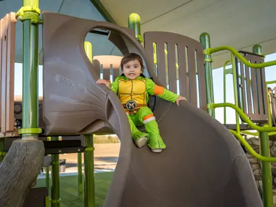 Picture of a young child dressed up as a Teenage Mutant Ninja Turtle sitting atop a brown slide at the new Waterleaf Playground in Kyle, TX. 
