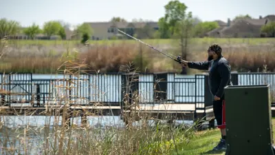 A man in a long-sleeved shirt wearing a hat and sunglasses casts a fishing pole near the dock at Lake Kyle Park in Kyle, TX.