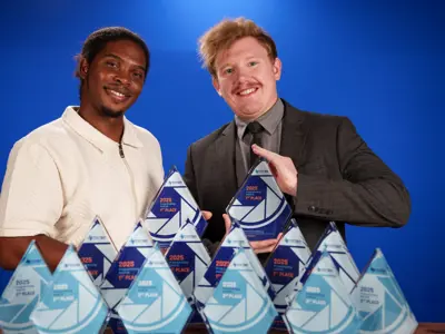 Two smiling men pose in front of a blue background, each holding a blue triangular award. Several similar awards are arranged on the table in front of them. One man wears a light shirt, and the other wears a suit.