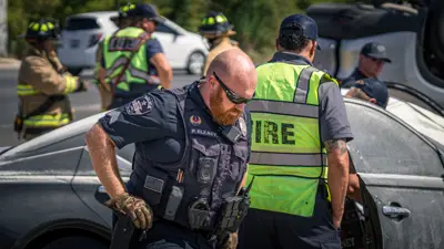 A Kyle Police Officer wearing sunglasses looks down on the ground as firefighters and EMS workers behind him tend to an automobile accident. 
