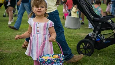 A young girl in a pink striped dress holds an Easter basket while wearing bunny ears at the Easter Egg-Stravaganza. 
