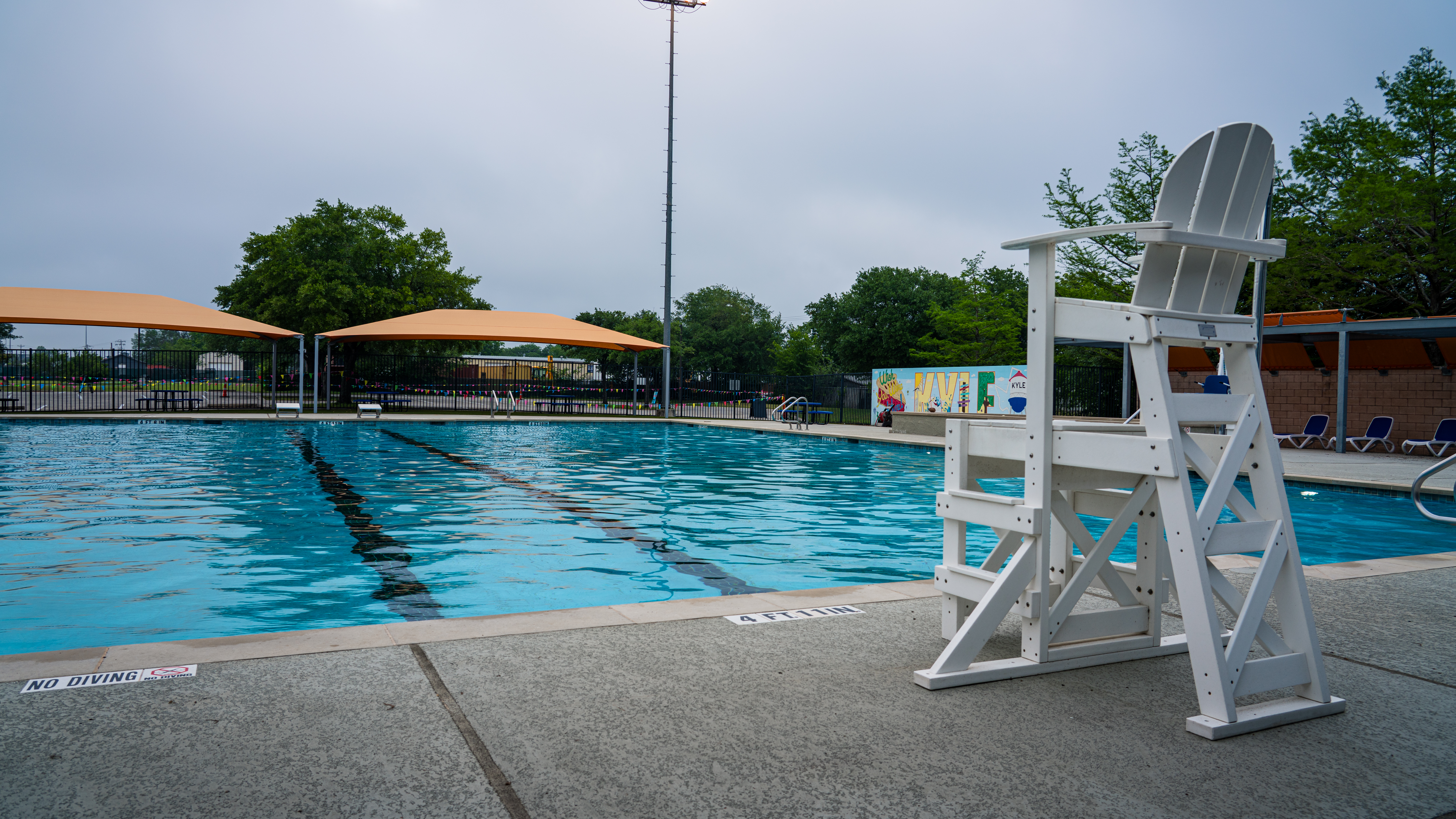 Picture of an empty James Adkin Pool in Kyle, TX. The blue water reflects off of a cloudy sky with a lifeguard chair in the foreground. 