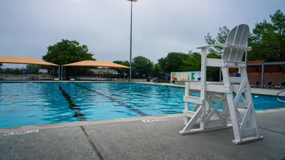Picture of an empty James Adkin Pool in Kyle, TX. The blue water reflects off of a cloudy sky with a lifeguard chair in the foreground.