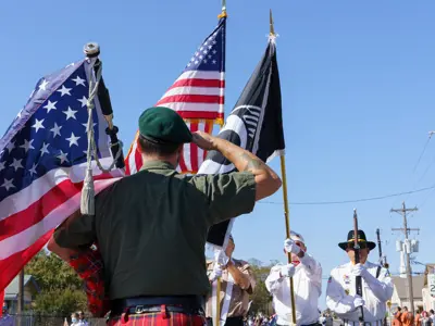 A man carrying a bag pipe salutes three Veterans dressed in white carrying the American and POW/MIA flags.
