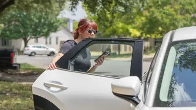 A young woman with reddish brown hair enters a white vehicle while holding her phone in her hand on a beautiful sunny day in Kyle, TX.