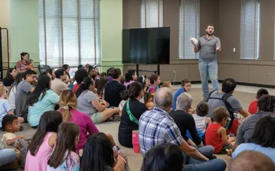 A man stands in front of a crowd sitting down showing a small animal to the crowd.