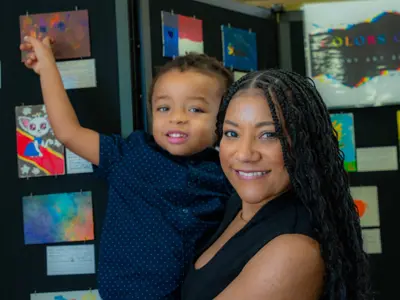 A young mom holds her son as he points to several pieces of art in the background as part of the Tiny Art Showcase at the Kyle Public Library. 